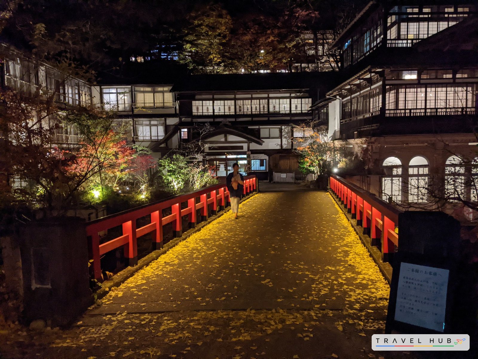 夜の四万温泉。銀杏の落ち葉が敷きつめられた赤い慶雲橋の向こうに、明かりのともる積善館本館が立つ